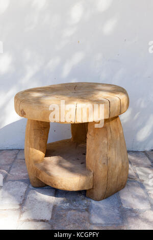 Round Sgabello in legno, realizzato a mano da un grande tronco di albero. Sorge su un pavimento di mattoni in prossimità di un edificio bianco. sulla superficie vi sono ombre dal crescente Foto Stock