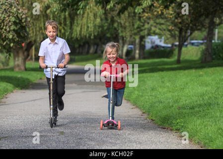 I bambini che cavalcano i loro scooter - un ragazzino e la sua giovane sorella che si divertono a correre lungo un percorso sugli scooter. Foto Stock