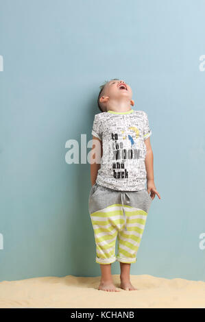 Little Boy in una maglietta e pantaloni corti in piedi sulla sabbia su sfondo azzurro. Foto Stock