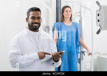 Ritratto di giovane maschio medico usando tavoletta digitale con la donna in fase di scansione a raggi x in background in ospedale Foto Stock