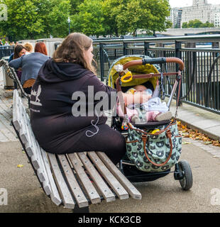 Il sovrappeso, genitore single, madre teenager, seduto su una panca in legno, allattamento al biberon il latte per il suo bambino in un passeggino, South Bank di Londra, Inghilterra, Regno Unito. Foto Stock