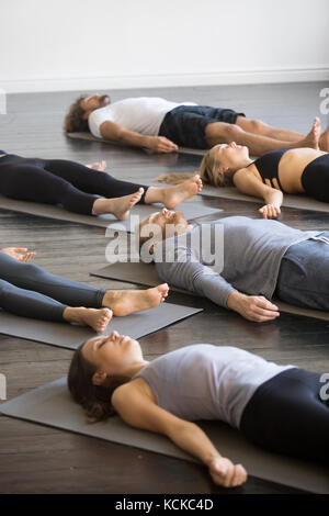 Un gruppo di giovani sportivi a praticare yoga lezione con istruttore in palestra, giacenti nel corpo morto pongono, facendo savasana cadavere, esercizio, amici rilassante Foto Stock