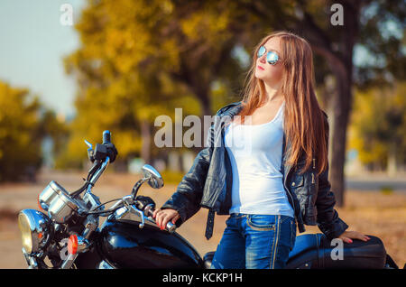 Elegante giovane donna in una giacca di pelle e jeans blu in un cavalletto di occhiali con una moto e e guarda su un urban autunno sfondo. Foto Stock