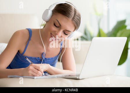 Sorridente ragazza teen indossando le cuffie ascolto corso audio utilizzando laptop a casa, rendendo note, giovane donna l apprendimento delle lingue straniere, digital sel Foto Stock