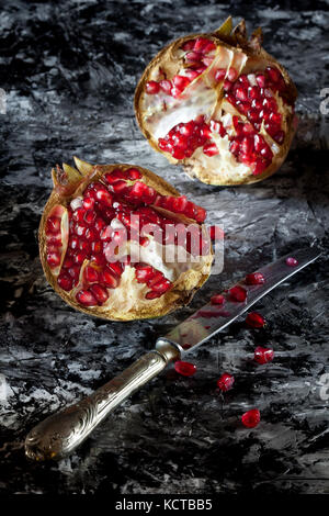 Melograno tagliato in due pezzi e argento coltello artistico su sfondo scuro. Foto Stock