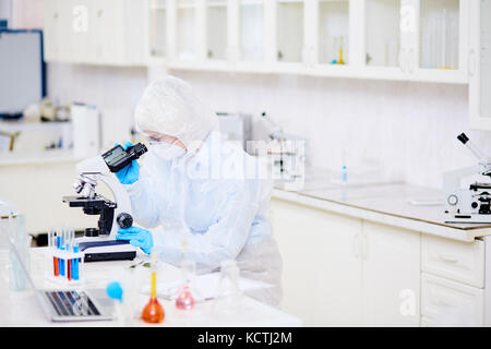 Vista di profilo di donna microbiologo indossando hazmat suit guardando attraverso il microscopio mentre studiava la mutazione virus al laboratorio moderno Foto Stock