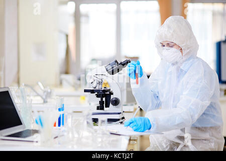 Microbiologa donna in tuta di nocciola che scrive i risultati di esperimenti condotti sugli appunti mentre si concentra sul lavoro in un laboratorio moderno Foto Stock