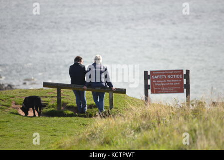 Due persone sedettero accanto a un cartello di sicurezza con il loro cane che guardava verso il mare in una soleggiata giornata autunnale, Filey, North Yorkshire UK Foto Stock