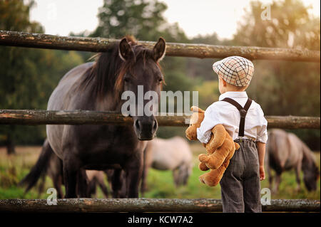 Un quattro-anno-vecchio ragazzo vestito in stile retrò è in piedi indietro tenendo un orsacchiotto e guardando un cavallo Foto Stock