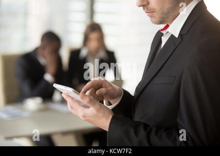 Chiudere l immagine dell uomo d affari maschiatura sul cellulare schermo con il dito, componendo il numero di messaggio di scrittura, l uomo e la donna in abbigliamento formale seduti alla scrivania mi Foto Stock