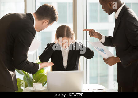 Sottolineato femmina di lavoratore di ufficio di chiudere le orecchie con le mani mentre è seduto alla scrivania circondata da mad nervoso colleghi o bugne rivolti a errori di rep Foto Stock