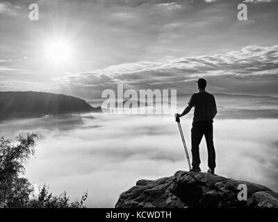 Tall guida turistica su roccia con la pole in mano. escursionista con equipaggiamento sportivo sopra misty valle soleggiata lo spuntar del giorno di primavera nelle montagne rocciose. Foto Stock