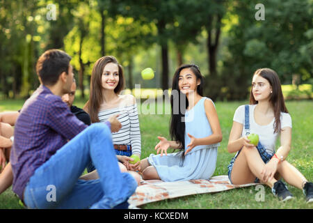 Amici su pic-nic, giovane uomo getta un apple per donna. Gli studenti aventi il divertimento a pranzo nel soleggiato parco. persone di diverse nazionalità trascorrono le loro fr Foto Stock