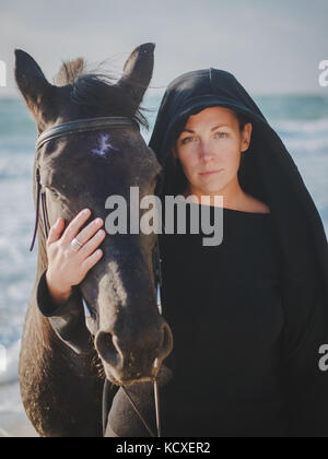 Giovane donna bionda stroking e abbracciando cavallo. bella signora con il suo stallone nero sulla riva del mare e la natura. amore e amicizia concetto onde. Foto Stock