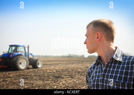 Giovane uomo in un campo e un trattore su uno sfondo. Concetto di agricoltura e di campo funziona. Foto Stock
