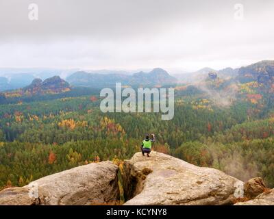 Hobby fotografo prende foto di primavera la natura parco da rocce appuntite. escursionista in giacca verde rimanere con la fotocamera sul cavalletto sul picco lapideo è aumentato da Foto Stock