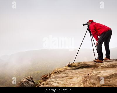 Escursionista con la fotocamera sul cavalletto prende foto dalla vetta rocciosa. fotografo solo in corrispondenza del bordo fotografia brumoso paesaggio, foresta in valle. Foto Stock