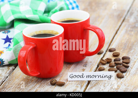 Buona mattina card con rosso di due tazze di caffè e i chicchi di caffè rustico superficie in legno Foto Stock