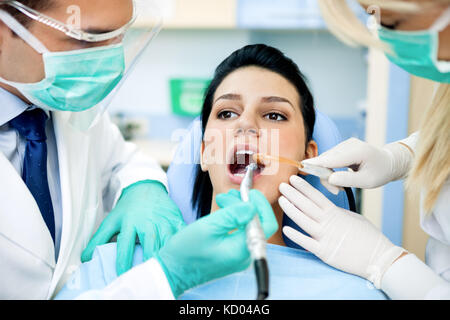 Paziente di sesso femminile con dentista e assistente, indossando maschere e guanti, dentista e assistente chinandovi sopra il suo utilizzo sucker e forare Foto Stock