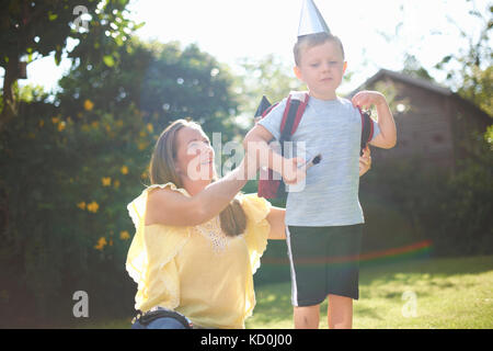 Donna matura mettendo a razzo zaino in costume sul figlio in giardino Foto Stock