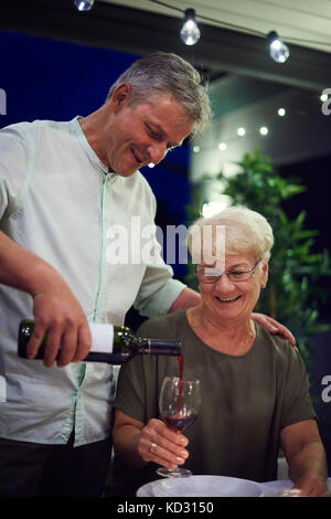 Senior donna seduta al tavolo per la cena, tenendo bicchiere da vino, uomo maturo versando il vino nel bicchiere Foto Stock