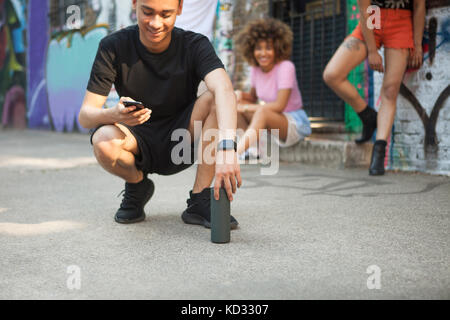 Tre amici appendere fuori strada, giovane uomo accovacciato, guardando a smartphone Foto Stock