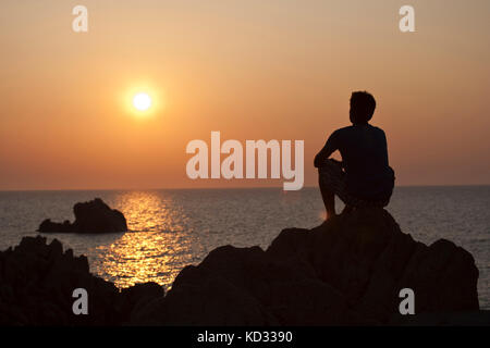 Silhouette di uomo sulle rocce che guarda lontano al tramonto sul mare, Olbia, Sardegna, Italia Foto Stock