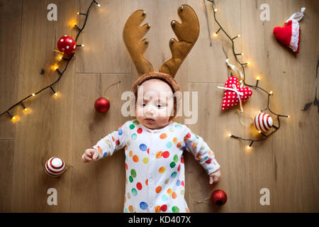 Little baby boy sdraiato sul pavimento che indossa la fascia di renne al tempo di Natale. ad alto angolo di visione. Foto Stock