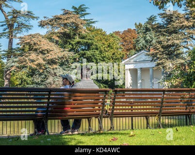 Asian giovane seduto su una panchina nel caloroso inizio autunno sole mattutino, accanto al tempio storico in Gunnersbury Park, West London, England, Regno Unito. Foto Stock