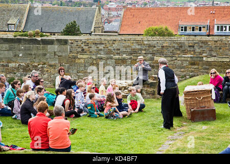 Bambini e adulti che ascoltano un Storyteller presso le rovine dell'abbazia sulla cima delle scogliere sopra Whitby Town raccontando una storia di Dracula Foto Stock