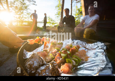 Immagine ritagliata della donna di mano la cottura alla griglia di cibi su spiedini con amici in background in foresta Foto Stock