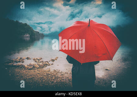 Donna con ombrello rosso contempla nella pioggia di fronte ad un lago. triste e solitario persona femmina cercando in distanza. grunge editing con sporcizia, rumore, Foto Stock