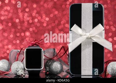 Smartphone e smartwatch e decorazioni per albero di Natale di vetro nero tabella su sfondo rosso. concentrarsi sullo smartphone. Foto Stock