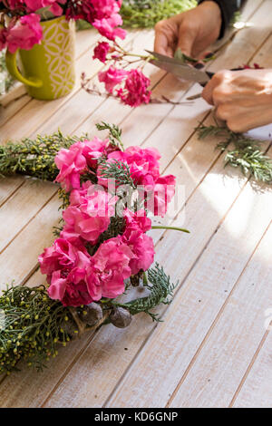 Fioraio al lavoro: creazione di una ghirlanda di legno con rami di pino e fiori di colore rosa e circondato dalle arti e mestieri. Foto Stock