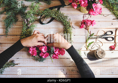 Fioraio al lavoro: creazione di una ghirlanda di legno con rami di pino e fiori di colore rosa e circondato dalle arti e mestieri. Tettuccio Foto Stock