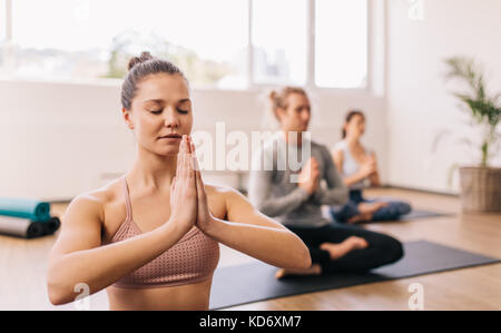 Giovane donna a praticare yoga in palestra classe nel centro fitness con persone di background. Persone meditando al club della salute. Foto Stock