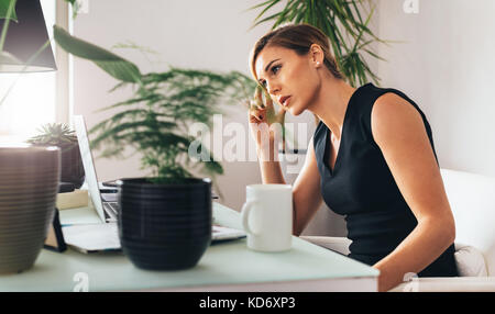 Malinconici cerca donna seduta alla sua scrivania in ufficio. Business donna guardando il computer portatile sul tavolo con la mano toccare la testa. Foto Stock