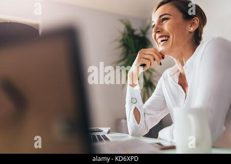 Donna sorridente seduto alla sua scrivania in ufficio. Happy business donna seduta in ufficio con dita toccano il suo mento. Foto Stock
