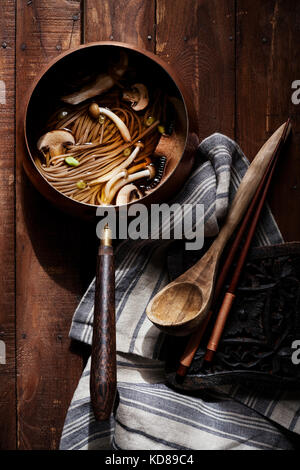 Pentola di rame del vecchio mondo piena di zuppa di noodle asiatiche e funghi su una superficie di legno ricca con cucchiaio di legno antico, lino e bastoncini di chopsticcio. Foto Stock