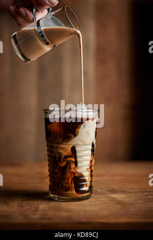 Bicchiere di caffè ghiacciato con creamer vintage versando in vetro sui toni caldi del legno. Foto Stock