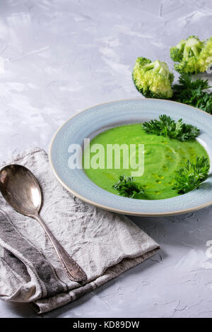 Vegetariano vegano crema di broccoli Zuppa servita nella piastra blu con prezzemolo fresco, broccoli, cucchiaio, tessile igienico oltre il cemento grigio Sfondo. Una sana e Foto Stock