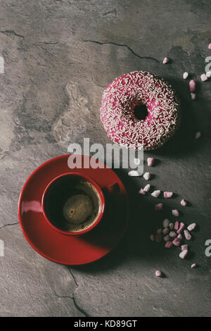 Vetri rosa ciambella con zucchero rosa e rosso tazza di caffè nero su grigio Sfondo texture. Laici piana con uno spazio Foto Stock