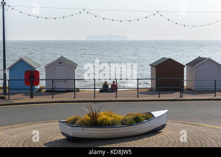 Coppia di spiaggia, vista posteriore di una coppia di mezza età seduta da sola tra capanne sulla spiaggia sul lungomare a Felixstowe, guardando una nave container distante, Regno Unito Foto Stock