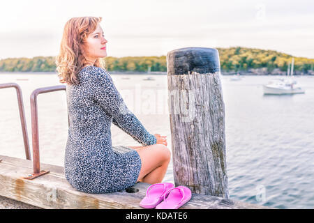 Giovani sorridenti donna seduta sul bordo del dock in Bar Harbor, Maine guardando acqua e bay con sole luminoso alla luce del sole, luce calda luce morbida Foto Stock