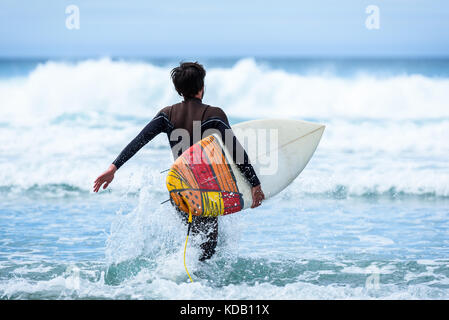 Surfer ragazzo con la tavola da surf in mano a correre verso grandi onde. l uomo nel surf muta umida è in esecuzione in ondate di freddo oceano atlantico in Galizia, Spagna. Foto Stock