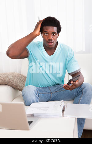 Ritratto di un preoccupato del giovane africano tenendo la calcolatrice di lavoro a casa Foto Stock