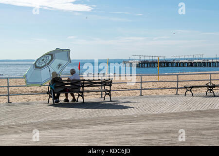 Una coppia di anziani seduti su una panchina sotto un ombrello su un caloroso inizio autunno mattina. In Coney Island Brooklyn, New York. Foto Stock