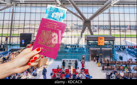 Mano che tiene il passaporto del Regno Unito e il Regno Unito 5 pound nota con segnale di occupato in attesa airport lounge e aeroplani in background Foto Stock