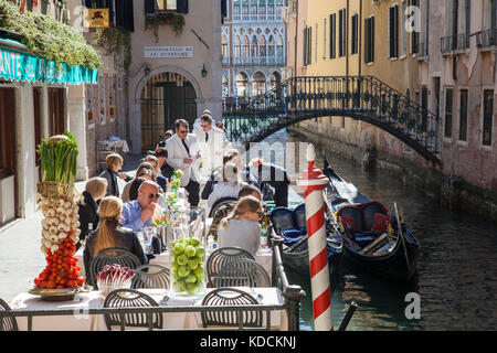 I turisti cenare presso il famoso Ristorante all'aperto da Raffaele, San Marco, Venezia, Italia al fianco di un canale con gondole e gondoliere in primavera sunsh