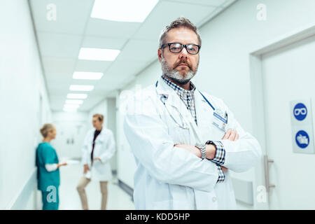 Ritratto di medico esperto in piedi nel corridoio di ospedale con le braccia incrociate. Caucasica medico maturo con i colleghi in piedi in background. Foto Stock